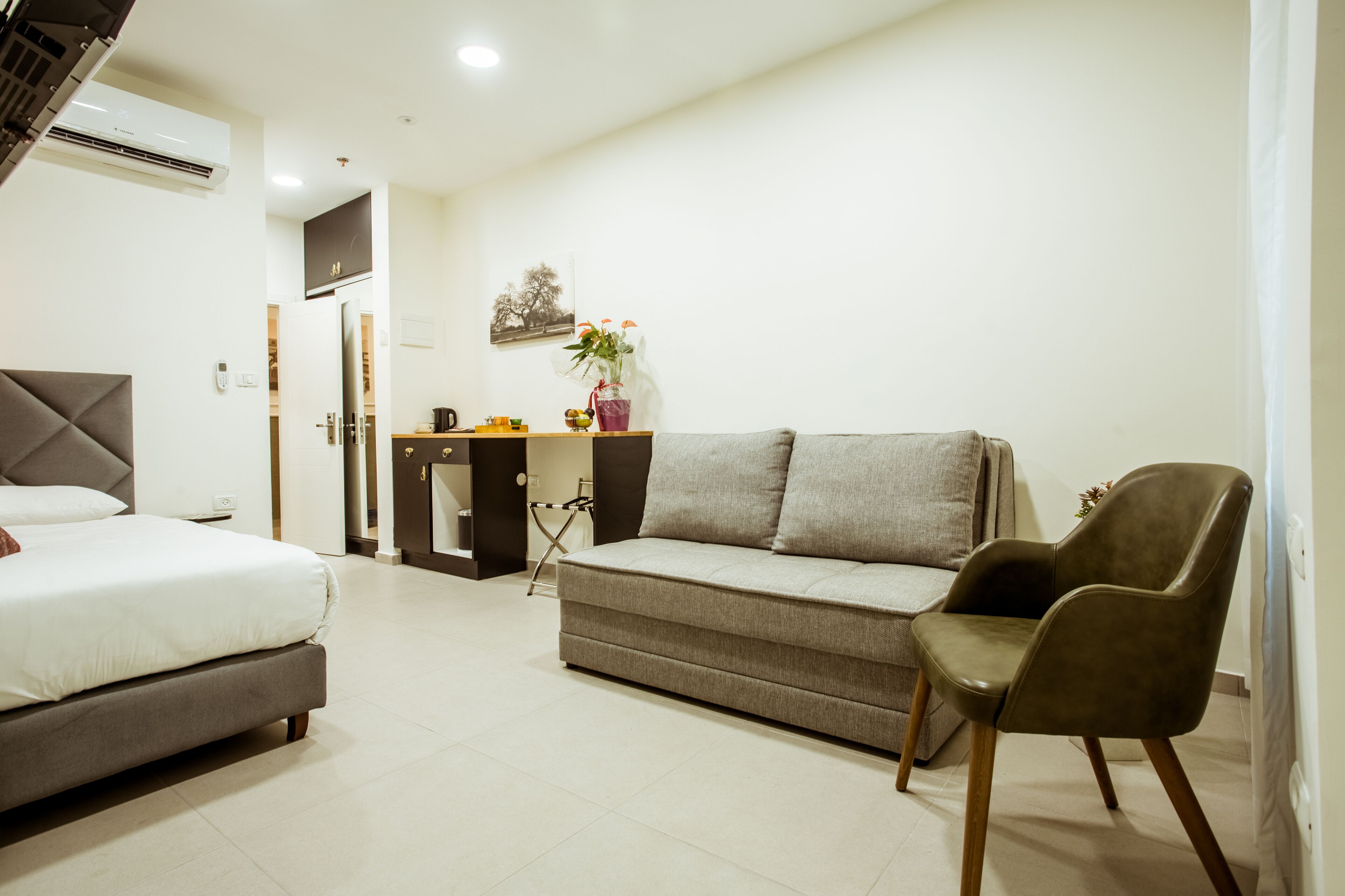 A modern hotel room with a bed, a sofa, a chair, a small desk, and a potted plant.