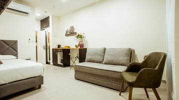 A modern hotel room with a bed, a sofa, a chair, a small desk, and a potted plant.