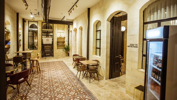 A spacious cafe with a patterned rug, wooden tables, and chairs. There is a meeting room sign on the wall and a refrigerator with beverages. - The Negev Hotel (Beersheba)