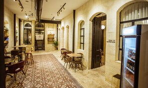 A spacious cafe with a patterned rug, wooden tables, and chairs. There is a meeting room sign on the wall and a refrigerator with beverages. - The Negev Hotel (Beersheba)