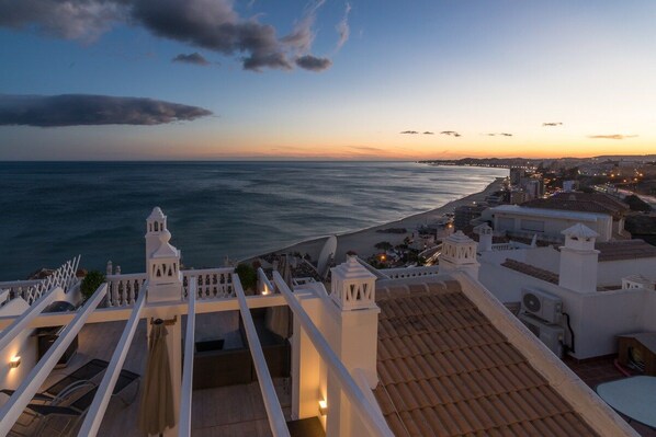 A rooftop terrace with a view of the sea and a cityscape at sunset.