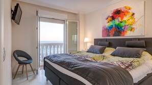 A modern bedroom with a large bed, a chair, a balcony, and a colorful abstract painting.