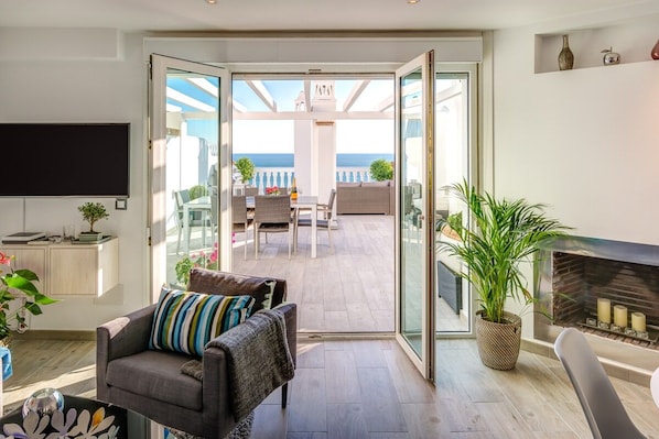 En stue med sofa, TV og pejs. Der er en balkon med havudsigt.