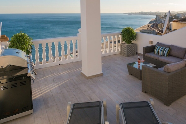 A balcony with a view of the sea, a grill, and seating area.