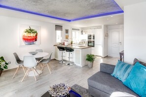 A modern living space with a dining area, kitchen, and a grey sofa with blue cushions.