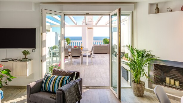 A modern living room with a sofa, a flat-screen TV, and a fireplace. There is a glass door leading to a balcony with a view of the sea.