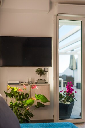 A room with a flat-screen TV mounted on the wall, a small cabinet with a plant, and a potted plant in the foreground.