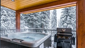 Un jacuzzi y una parrilla en una terraza de madera con vista a un bosque nevado.