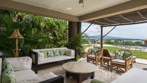 Un vaste espace extérieur aménagé avec un canapé, des fauteuils et une table basse, entouré d’une végétation luxuriante et offrant une vue sur une piscine et des montagnes.