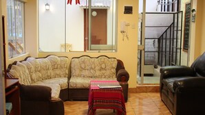 A living room with a sofa, a small table with a tablecloth, a leather chair, and a staircase visible through a glass door. - Casa de Ana Huaraz (Independencia)