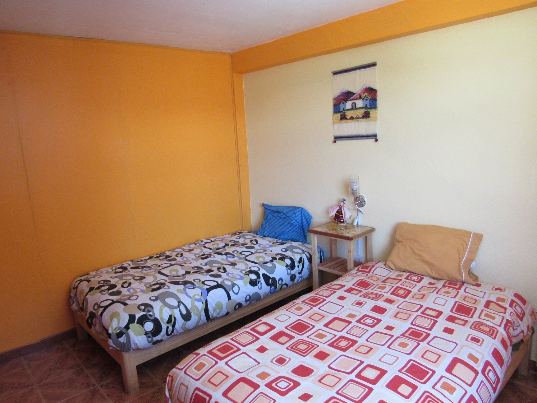 A room with two beds, one with a patterned bedspread and the other with a geometric design. There is a small table with a lamp and a decorative item on it. The walls are painted yellow, and there is a painting on the wall.