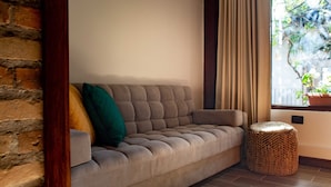 Un canapé avec un pouf en osier à côté, une fenêtre avec des rideaux et un mur en briques.