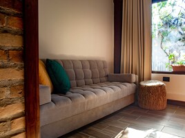 A sofa with a woven ottoman beside it, a window with curtains, and a brick wall.