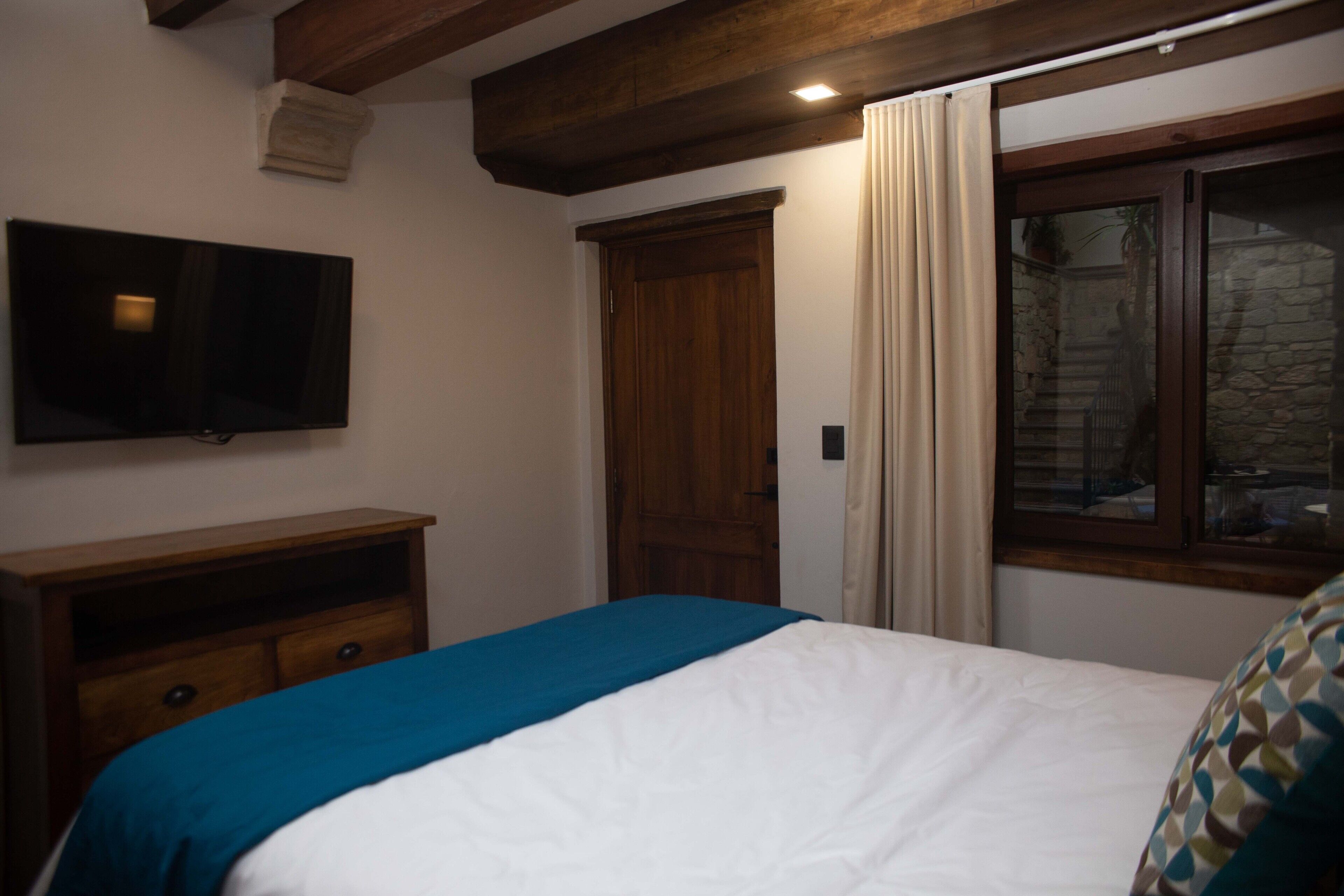 Un dormitorio con cama, cómoda de madera, televisor montado en la pared y ventana con cortinas.