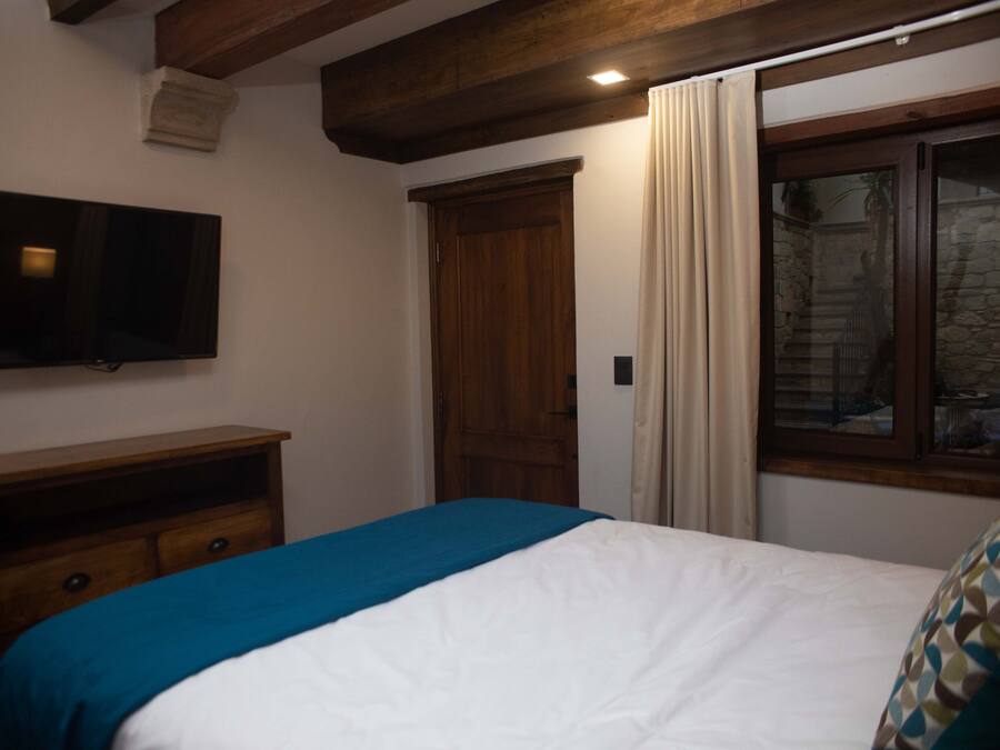 Un dormitorio con cama, cómoda de madera, televisor montado en la pared y ventana con cortinas.