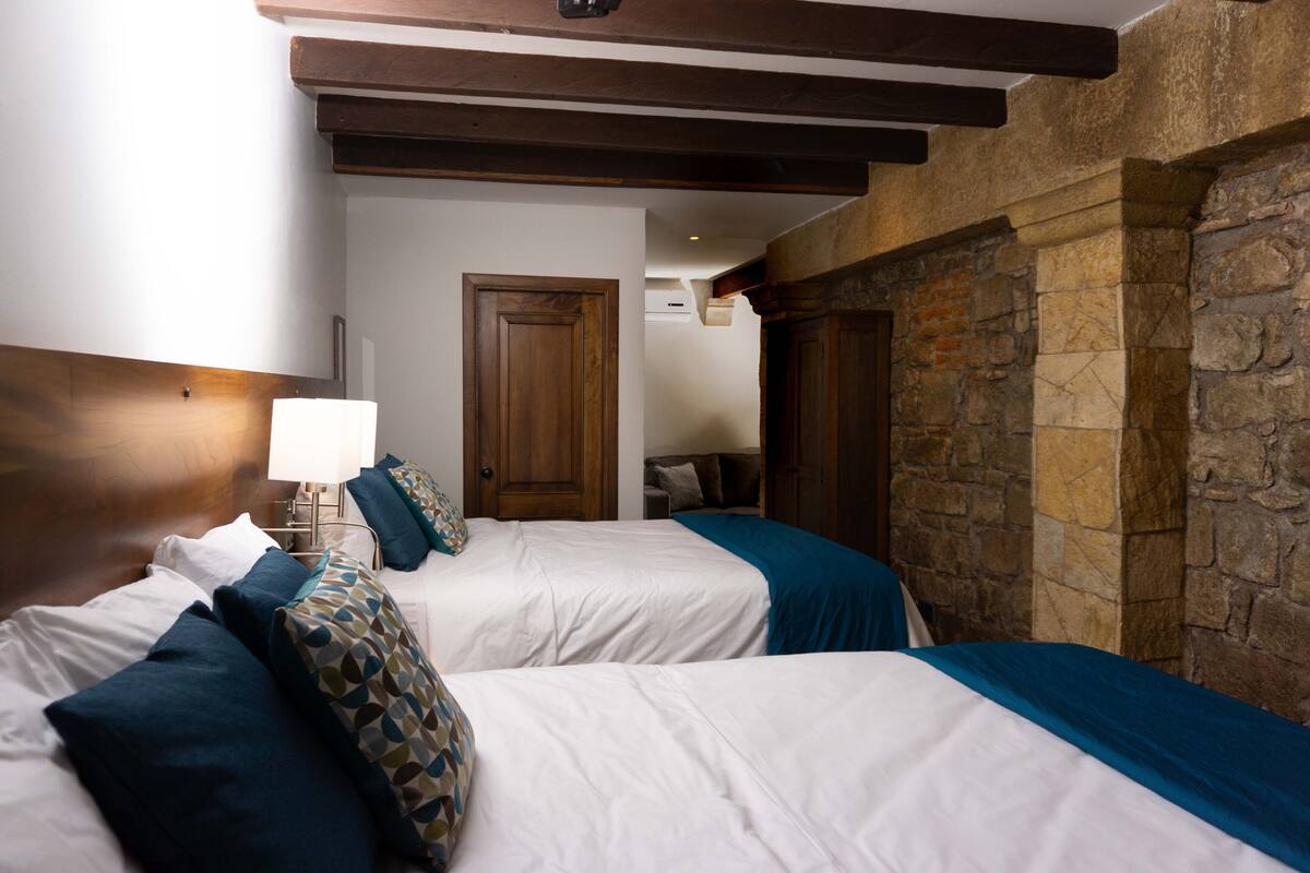 Ein Hotelzimmer mit zwei Betten, Holz-Kopfteilen, Steinwänden und einer Tür zu einem anderen Raum.