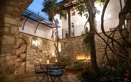 A stone courtyard with a table and chairs, surrounded by plants and a pergola.