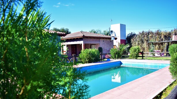 A swimming pool with a tiled deck, surrounded by greenery and a building with a tiled roof. - Cabañas Villa del Sol (Rosario de Lerma)