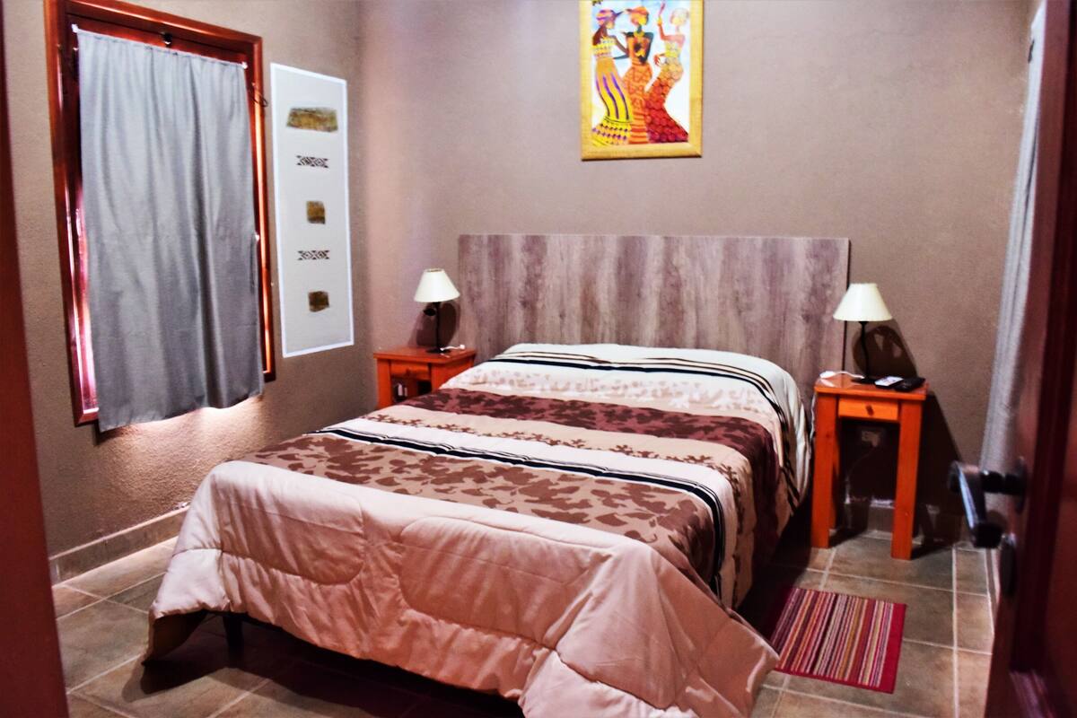 Habitación de hotel con una cama grande, dos mesitas de noche, una obra de arte colgada en la pared y una puerta con cortina.