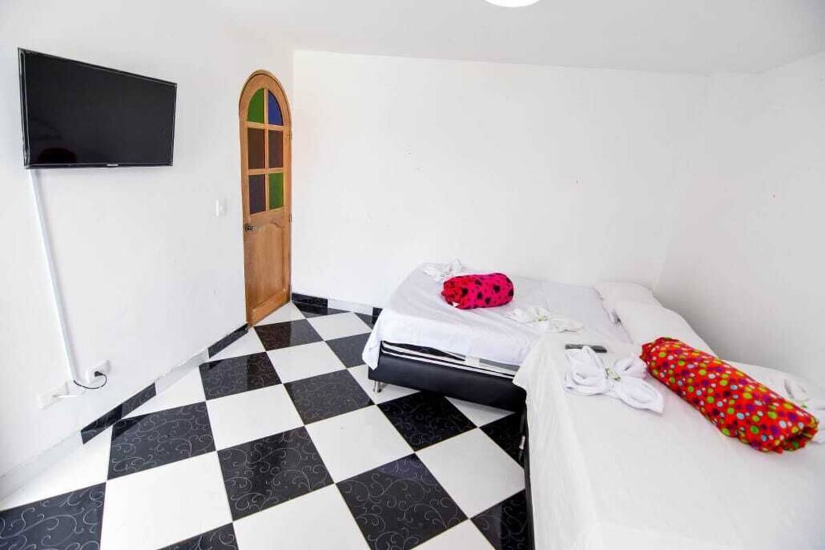 A room with a checkered floor, a bed with a white and red pillow, a TV mounted on the wall, and a wooden door with a colorful window.