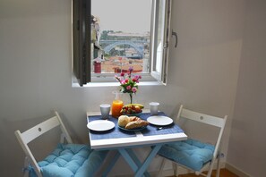 A small dining area with a table set for two, a vase with flowers, and a view of a bridge and buildings.