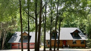 A two-story house with a red garage door, surrounded by tall trees and a gravel driveway. - Cedarwood Lake House (Toccoa)