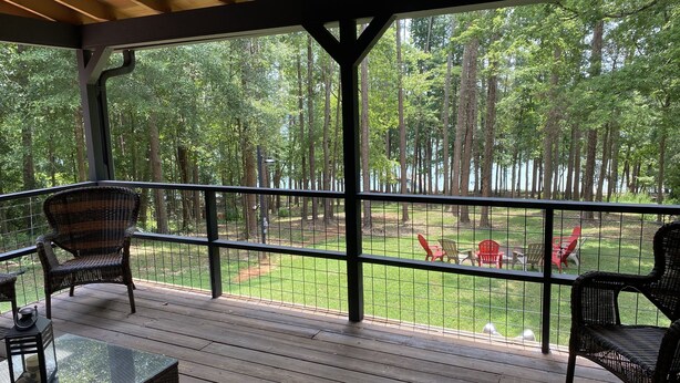 A covered deck with wicker furniture and a glass table, overlooking a grassy area with trees and a body of water. - Cedarwood Lake House (Toccoa)