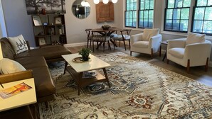 A living room with a brown sofa, white armchairs, a coffee table, a patterned rug, a dining table with chairs, and a large window with a view of trees. - Cedarwood Lake House (Toccoa)