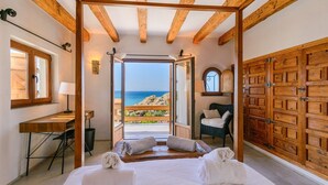 A bedroom with a wooden ceiling, a desk, a chair, a bed with towels, and a view of the sea.