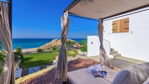A balcony with a view of the sea, a white wall with a window, and a wooden deck.