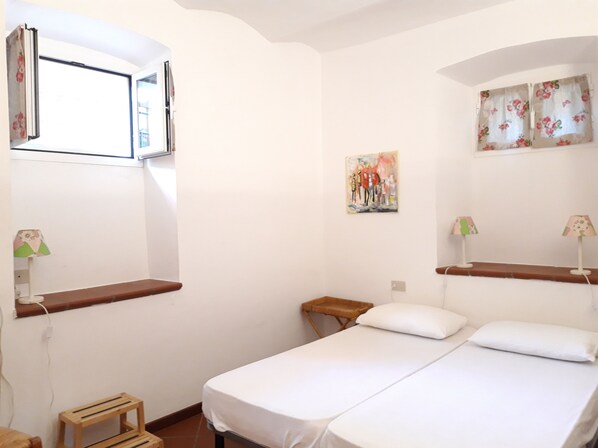 A bedroom with two beds, a window, a painting, and a small wooden stool.