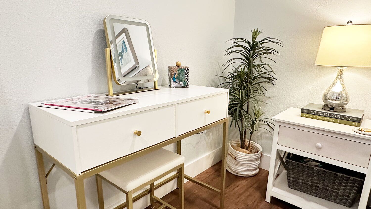 A white desk with a picture frame, a small potted plant, a lamp, and a book titled 'INTERIOR DECORATION' on a nightstand.