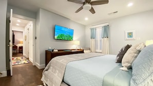 Un dormitorio con cama, ventilador de techo, televisor en la pared y ventana con cortinas.