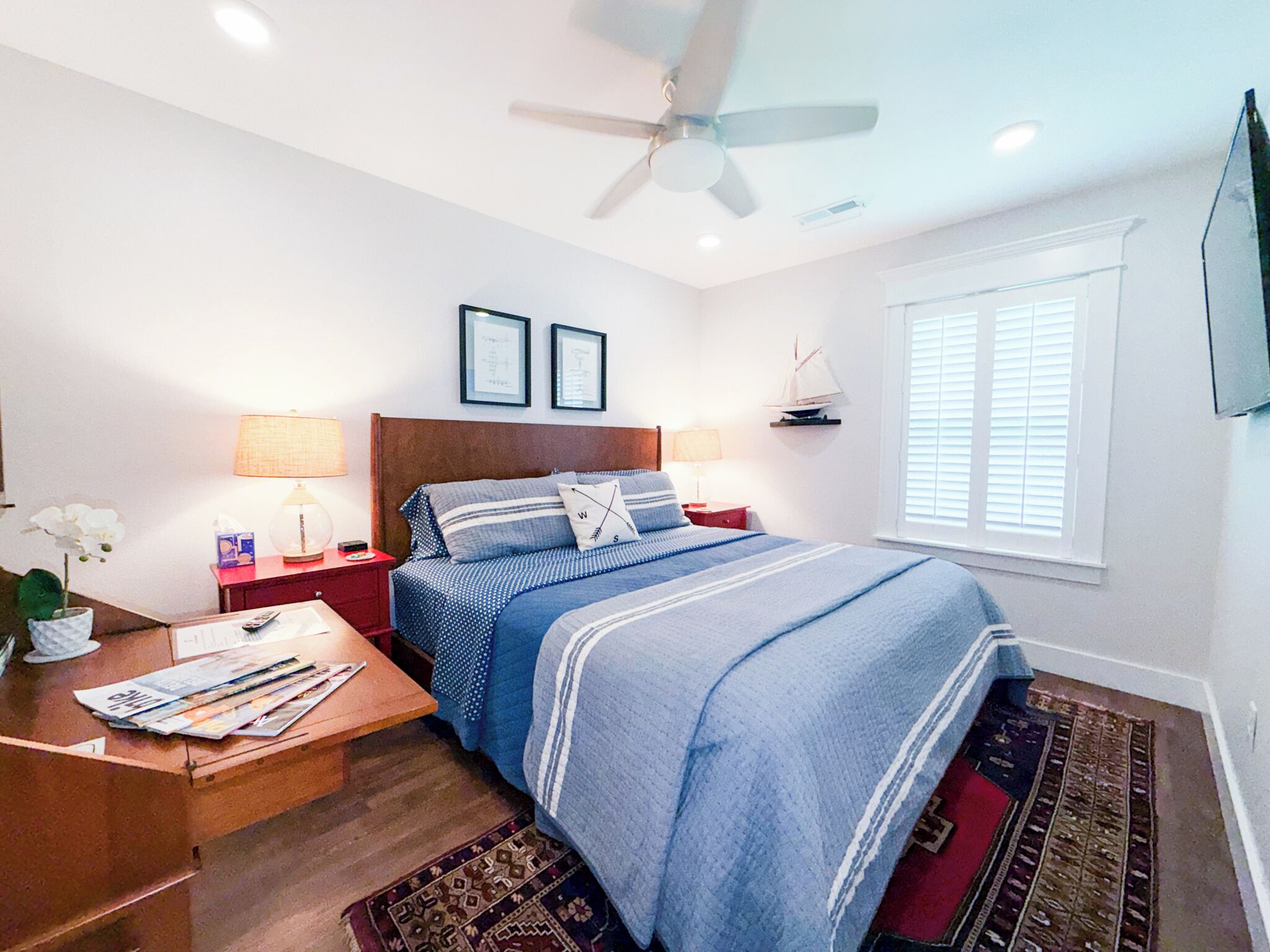 A bedroom with a bed, nightstands, a ceiling fan, a window with blinds, and a small sailboat decoration.