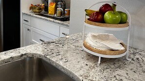 Una cocina con armarios blancos, un lavamanos de acero inoxidable, un microondas y una exposición de frutas.