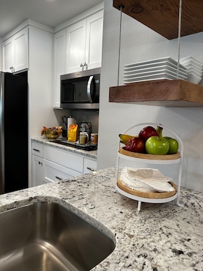 Una cocina con armarios blancos, un lavamanos de acero inoxidable, un microondas y una exposición de frutas.