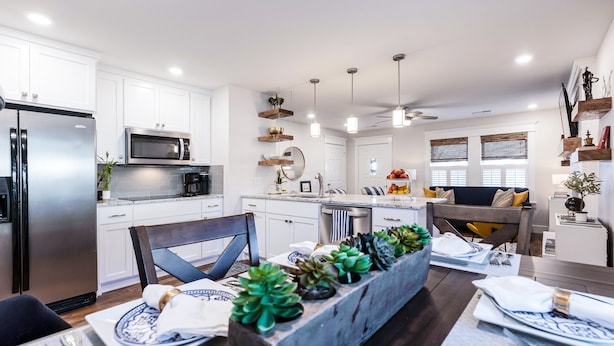 Una cocina moderna con armarios blancos, electrodomésticos de acero inoxidable y un comedor con una mesa de madera y plantas.