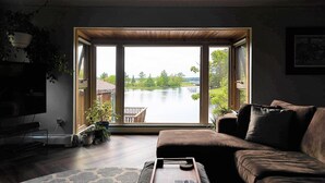 Un salon doté d’une grande porte-fenêtre coulissante donnant sur un lac, d’un canapé en cuir marron, d’une petite table et d’un téléviseur placé à gauche.