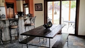 Una cocina rústica con una mesa de madera y bancos, un lámpara colgante, y vista a un área exterior por un ventanal.