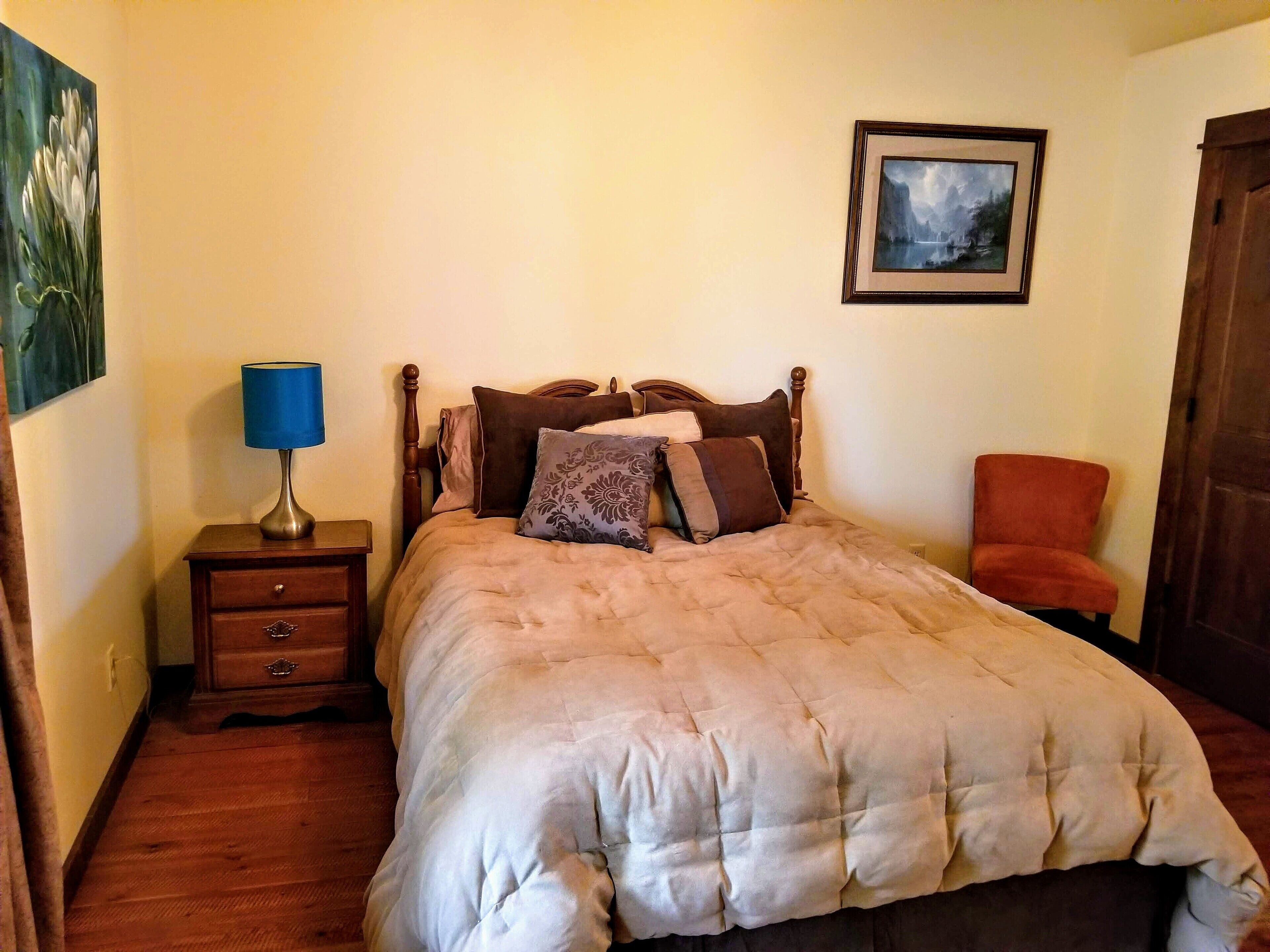 Une chambre à coucher avec un lit, une table de chevet, une lampe, une chaise et un tableau encadré au mur.