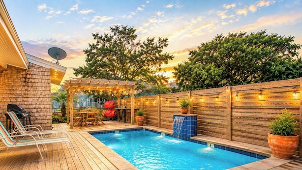 Un jardin avec une piscine, des chaises longues, un barbecue et une pergola illuminée de guirlandes lumineuses.