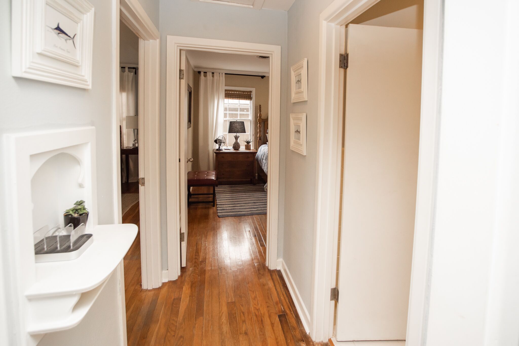 A hallway with wooden flooring, white walls, and a door leading to a room with a window and a small table.