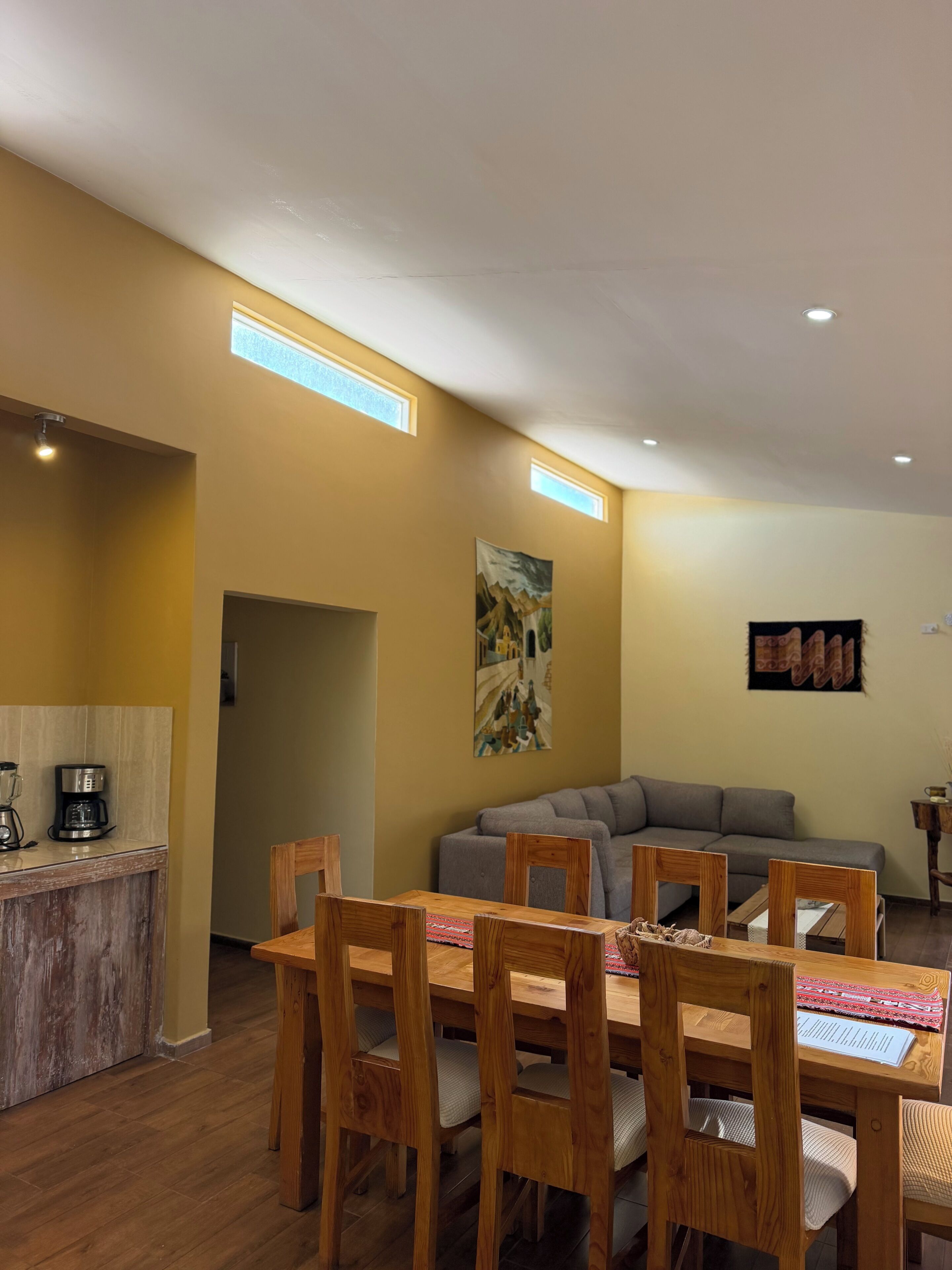 A dining area with wooden chairs and a table, a gray sofa, and a kitchen area with a coffee maker.