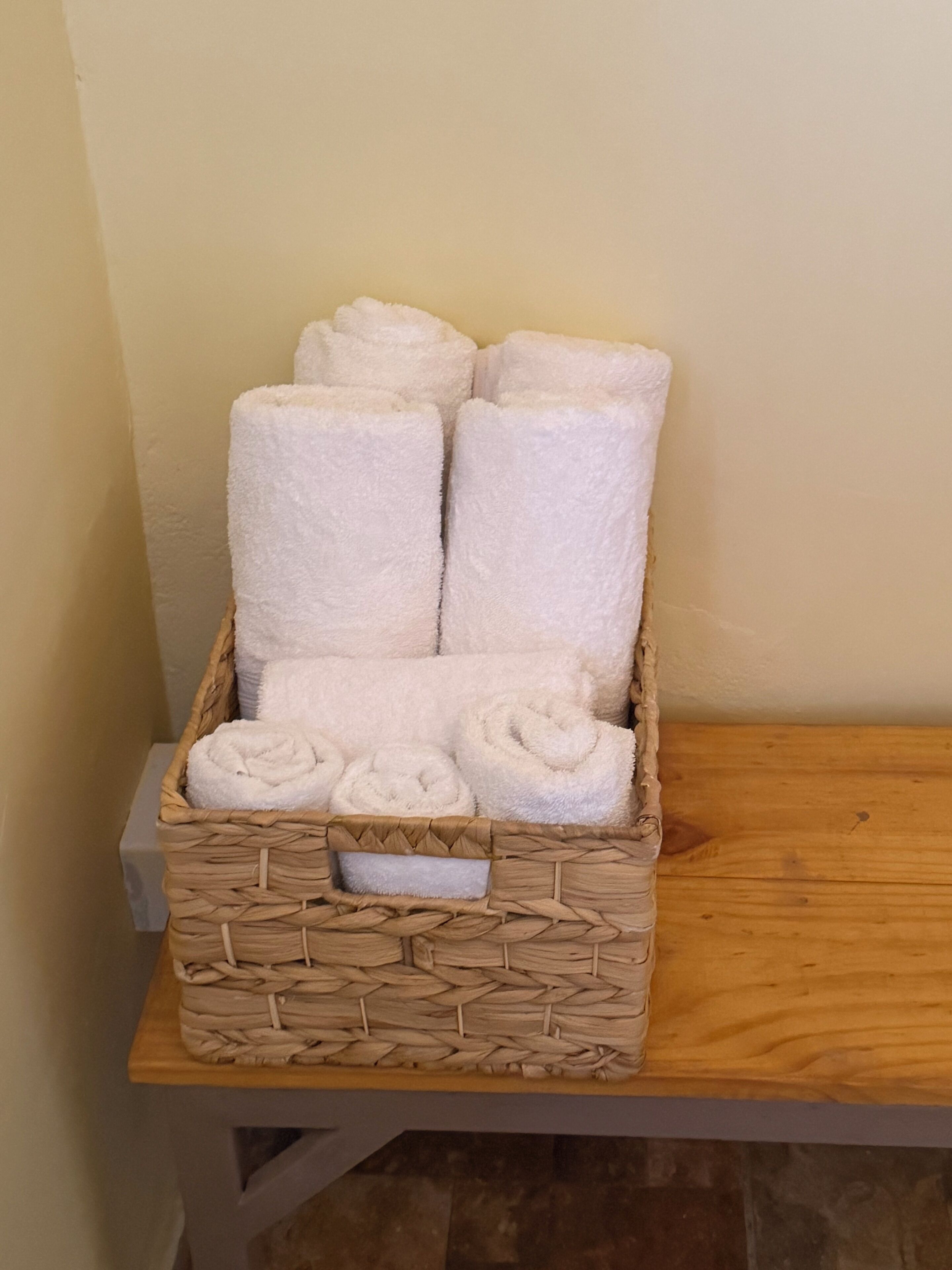 A wicker basket containing neatly rolled white towels on a wooden shelf.