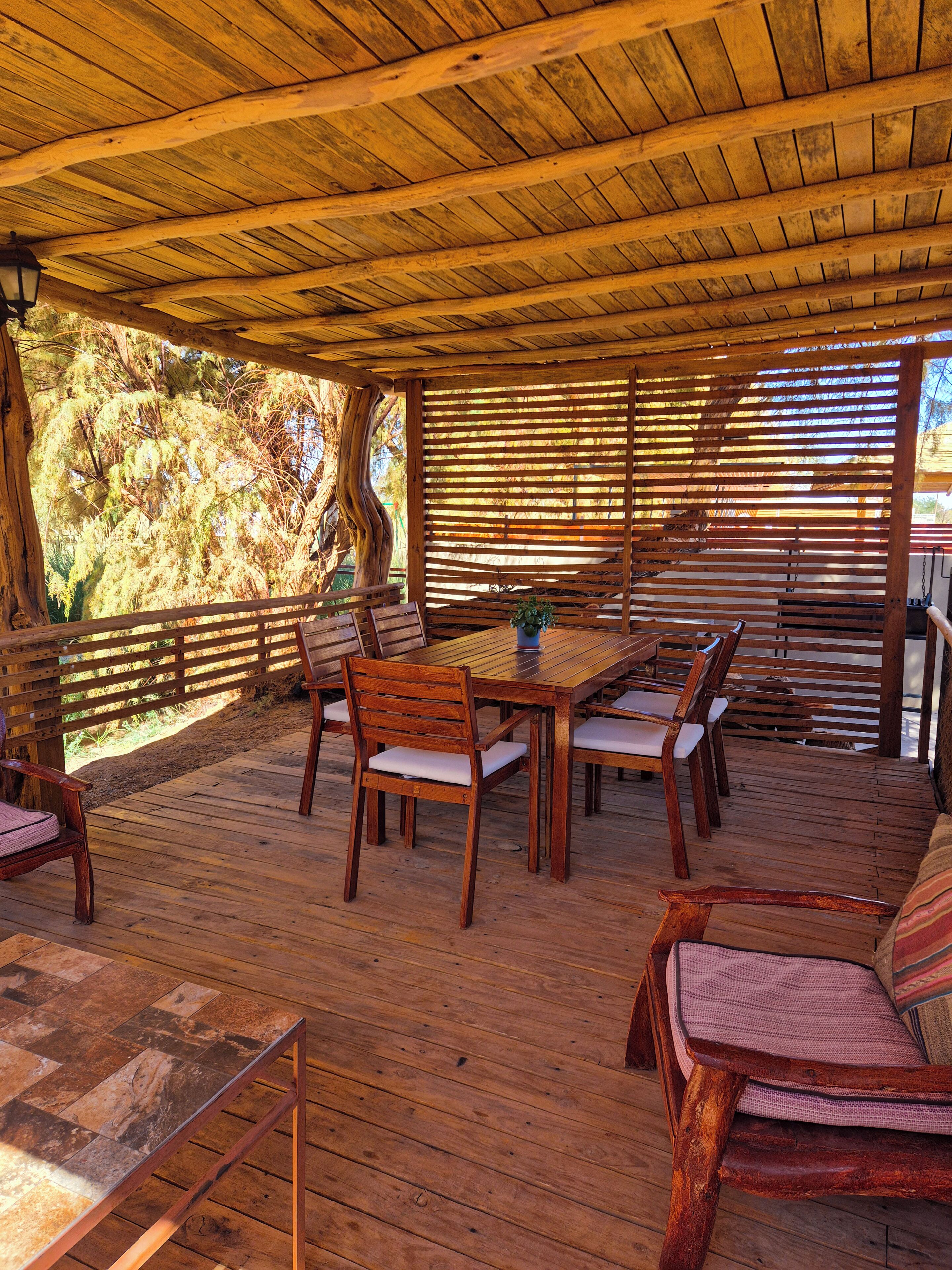 An outdoor wooden deck with a table and chairs, surrounded by wooden railings and a view of trees.