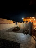 Un espacio exterior nocturno con un sendero pavimentado, un pequeño jardín con plantas y un edificio de fachada de madera.