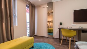A modern hotel room with a yellow sofa, a blue rug, a desk with a chair, and a TV on the wall. - Vanilla Boutique Hotel (Skopje)