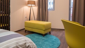 A modern bedroom with a bed, a yellow armchair, a small sofa, a standing lamp, and an air conditioning unit. - Vanilla Boutique Hotel (Skopje)
