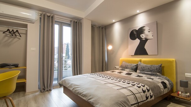 A modern bedroom with a large bed, a yellow chair, and a city view through the window. - Vanilla Boutique Hotel (Skopje)