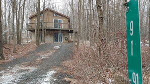A cabin with a balcony, surrounded by trees and a gravel path leading to it.